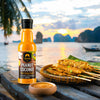 SIAM Peanut Coconut Thai Satay Sauce bottle with satay skewers and dipping sauce on a wooden platform by a scenic waterfront.