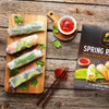 Spring rolls on a wooden board with a small bowl of sauce, a pair of chopsticks, and a SIAM Spring Roll package on a rustic wooden surface.