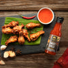 Fried chicken wings on a banana leaf with a bottle of Siam Sweet Chilli Hot Sauce on a wooden surface.