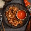 Plated dish of spicy chicken with a side of rice and a bottle of spicy sauce on a wooden table.